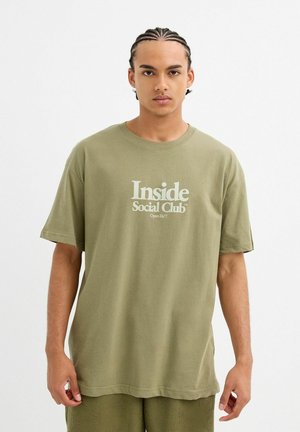 Camiseta de algodón verde oliva con mangas cortas, que presenta un logo blanco de "Inside Social Club" impreso en el frente. Corte casual con escote redondeado.