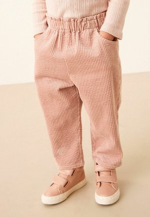 Enfant portant un pantalon côtelé rose avec taille élastique, mains dans les poches, et des baskets roses assorties à scratch, debout sur un sol beige.