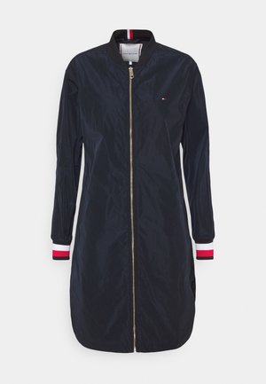 Long navy bomber jacket with front gold zipper, ribbed collar and cuffs featuring red, white, and black stripes, small chest logo.