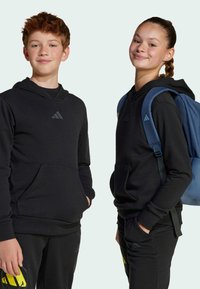 Felpe con cappuccio nere dotate di tasche a marsupio e logo. Un modello indossa uno zaino blu. Entrambi indossano pantaloni sportivi neri. Texture del tessuto morbida.