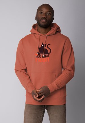 Roestkleurige hoodie met een kangoeroezak en een capuchon met trekkoord, voorzien van een grafische afbeelding van een zwarte kat en de zin "IK DOE WAT IK WIL" in oranje.