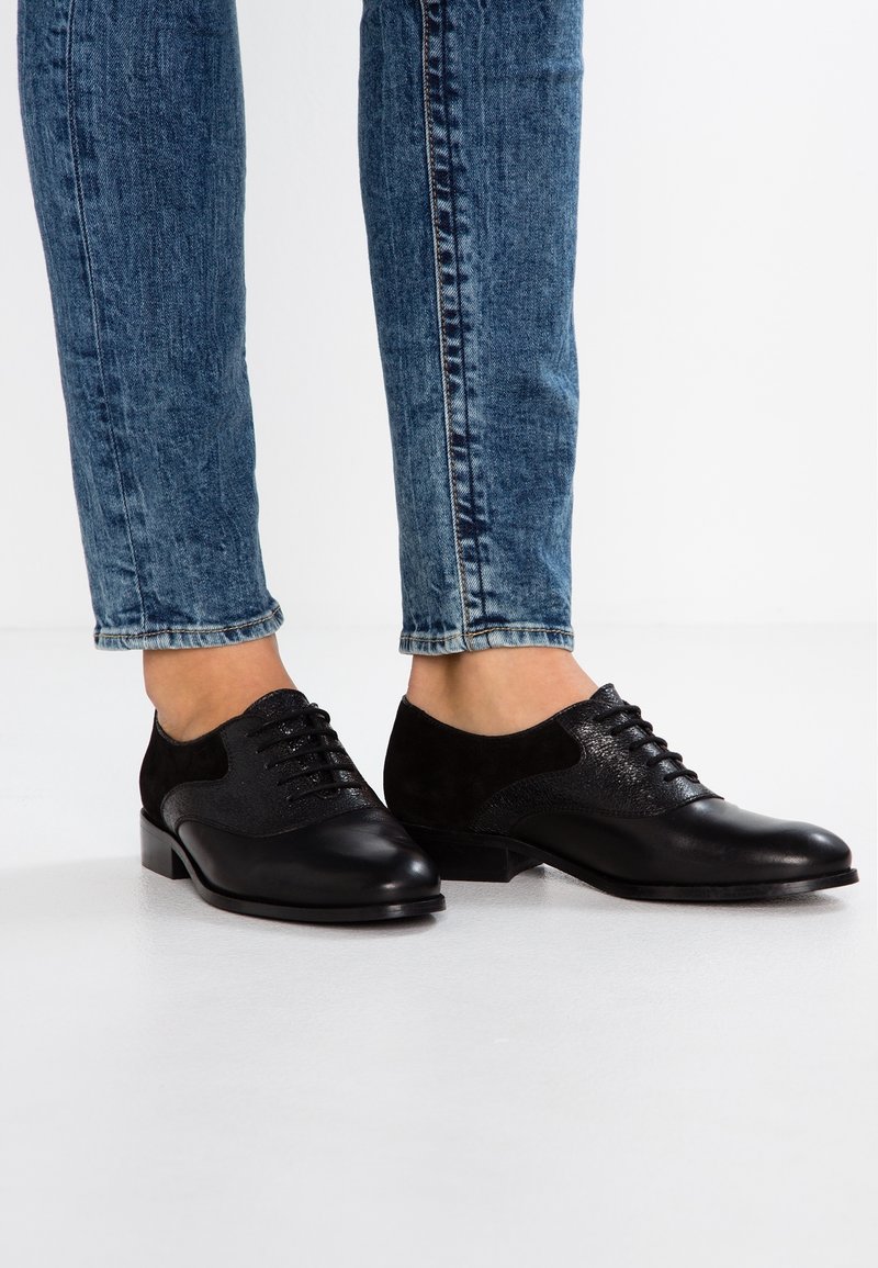 Pieds portant des chaussures Oxford en cuir noir à lacets associés à un jean en denim bleu, debout sur un sol blanc.