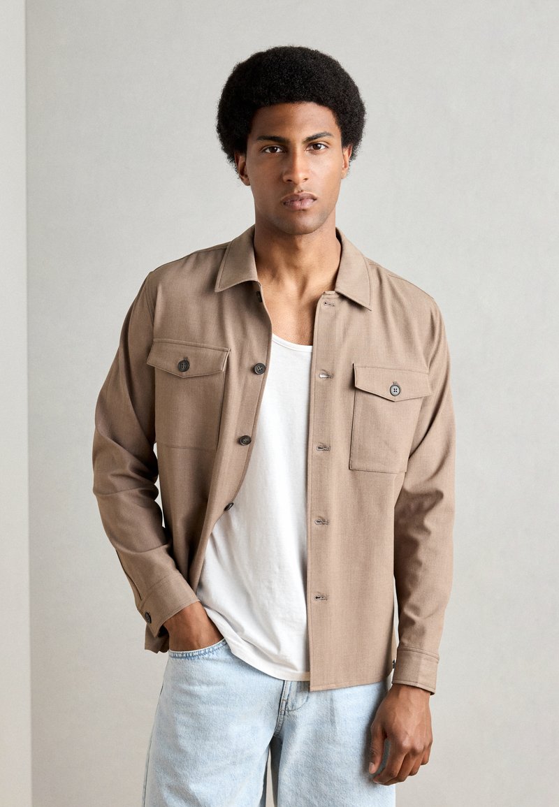 Cămașă button-up de culoare marron deschis, cu două buzunare pe piept, guler și croială lejeră, combinată cu un tricou alb și blugi de culoare albastru deschis.
