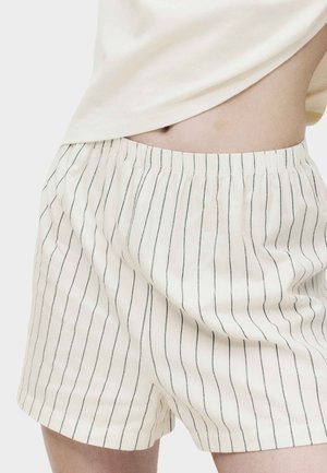 Persona con pantalones cortos blancos y sueltos con finas rayas negras verticales y una camiseta beige clara levantada para mostrar la cintura y el ombligo.