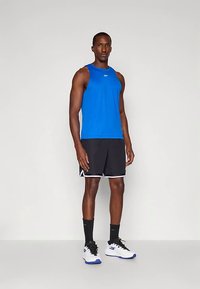 Top deportivo sin mangas de color azul, pantalones cortos negros con ribetes blancos y zapatillas blancas con acentos azules. El modelo está de pie sobre un fondo neutro.