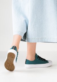 Baskets en toile bleu sarcelle avec des lacets noirs et des semelles blanches, présentant des panneaux latéraux texturés et une semelle extérieure en caoutchouc gomme.