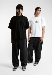 Two models wear oversized t-shirts, one black with a logo, the other white with a detailed graphic. Both style loose black pants and sneakers.