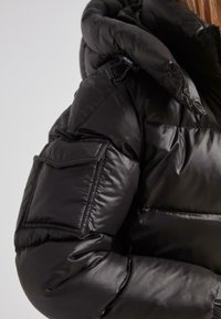 Acercamiento a una chaqueta de plumas negra y brillante con un gran bolsillo acolchado en la manga y un cordón cerca del hombro.