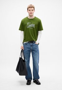 Young man in green Calvin Klein T-shirt over white long sleeves, blue jeans, black shoes, holding a large black leather tote bag.