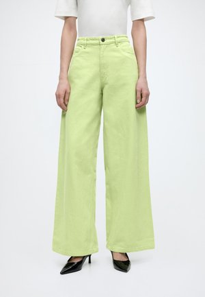 Person iført limegrønne bukser med brede ben, hvid kortærmet top og sorte spidse høje hæle, stående mod en ensfarvet baggrund.