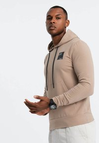 Felpa beige con patch logo nero, maniche lunghe, coulisse regolabile e tasca frontale. Il modello indossa un orologio nero al polso.