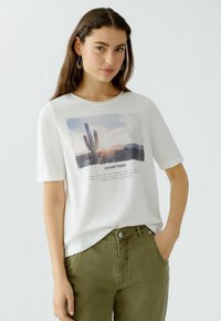 Weißes Baumwoll-T-Shirt mit einer Grafik eines Kaktus und Sonnenuntergang, mit kurzen Ärmeln und rundem Halsausschnitt, kombiniert mit olivgrünen Hosen.