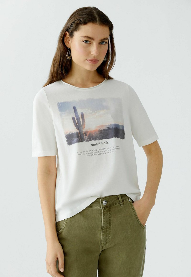 Weißes Baumwoll-T-Shirt mit einer Grafik eines Kaktus und Sonnenuntergang, mit kurzen Ärmeln und rundem Halsausschnitt, kombiniert mit olivgrünen Hosen.