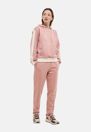 Femme en sweat à capuche rose avec des bandes blanches sur les manches et un pantalon assorti, portant des baskets beiges et marron, debout les mains dans les poches.