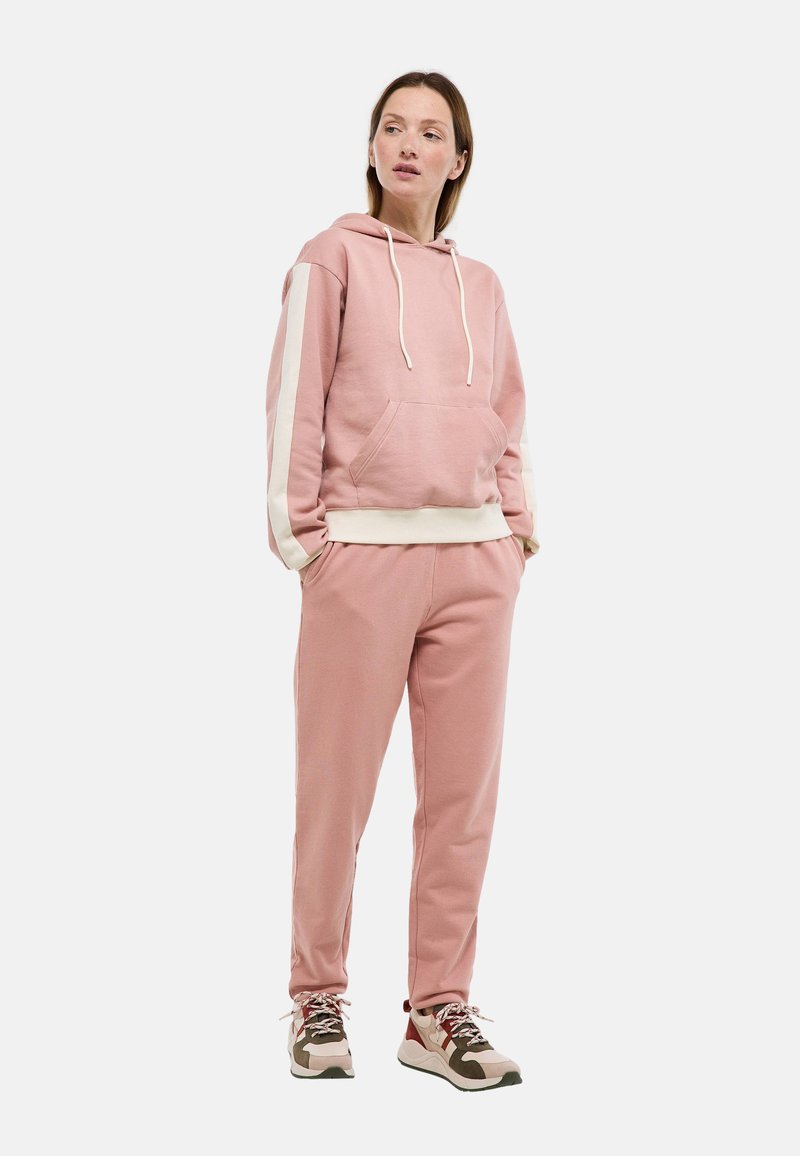 Femme en sweat à capuche rose avec des bandes blanches sur les manches et un pantalon assorti, portant des baskets beiges et marron, debout les mains dans les poches.