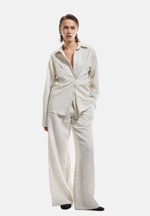 Outfit leggero e strutturato in due pezzi, composto da una camicia avvolgente con nodo davanti e pantaloni a gamba larga, entrambi color crema con motivi delicati.