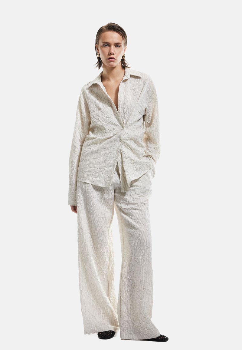 Leichtes, strukturiertes Zweiteiler-Outfit mit einem Wickelshirt mit vorderem Knoten und weit geschnittenen Hosen, beide in Creme mit dezenten Mustern.