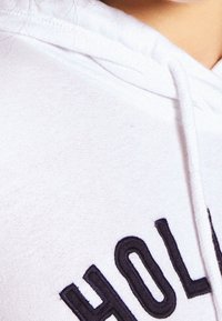 Vit hoodie med mjuk textur, med marinblå broderade bokstäver som stavar "HOLL." Inkluderar en dragkedja huva.