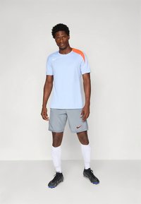 Světle modré sportovní tričko s oranžovým akcentem, doplněné šedými šortkami s oranžovým logem Nike a bílými knižními ponožkami, nošeno s černými kopačkami.