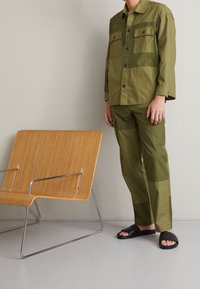 Completo due pezzi verde oliva con camicia con bottoni e pantaloni in stile cargo. Presenta tasche a toppa e sandali neri scivolati. Sedia di legno sullo sfondo.