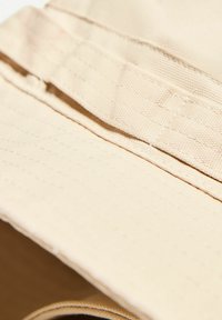 Beżowa czapka typu bucket wykonana z teksturowanej tkaniny. Posiada szeroki rondo i przeszycia wzdłuż korony, które dodają jej struktury.