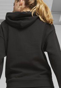 Čierna mikina s kapucňou vyrobená z mäkkej tkaniny, s veľkou kapucňou, voľným strihom a rebrovanými manžetami a lemom. Minimalistický dizajn bez viditeľných vzorov.