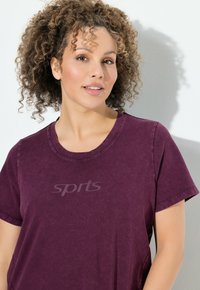 Lila Baumwoll-T-Shirt mit rundem Ausschnitt und kurzen Ärmeln. Vorne befindet sich ein dezentes "Sports"-Logo. Weicher Stoff, lässiges Design.