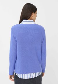 Pull en maille violet clair à manches longues, texture côtelée et ourlet arrondi, superposé à un haut à colrayé blanc et bleu.