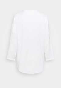 Camicia bianca a maniche lunghe con scollatura rotonda e vestibilità ampia. La texture sembra morbida; l'orlo è leggermente curvato nella parte inferiore.