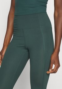 Dunkelgrüne, hoch geschnittene Leggings aus dehnbarem Stoff mit Flatlock-Nähten und Seitenpaneelen. Glatte Textur, figurschmeichelndes Design.