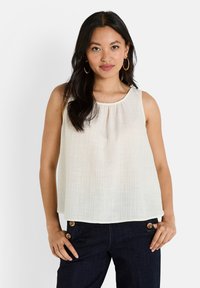 Haut sans manches, couleur blanc cassé, en tissu léger et texturé avec un col rond et des détails froncés. Associé à un pantalon en denim foncé avec des boutons dorés.