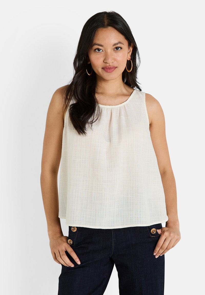 Haut sans manches, couleur blanc cassé, en tissu léger et texturé avec un col rond et des détails froncés. Associé à un pantalon en denim foncé avec des boutons dorés.