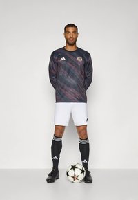 Camisola desportiva de manga comprida com padrão escuro com redemoinhos vermelhos e azuis, calções brancos, meias pretas com riscas brancas e sapatos de futebol pretos, segurando uma bola.