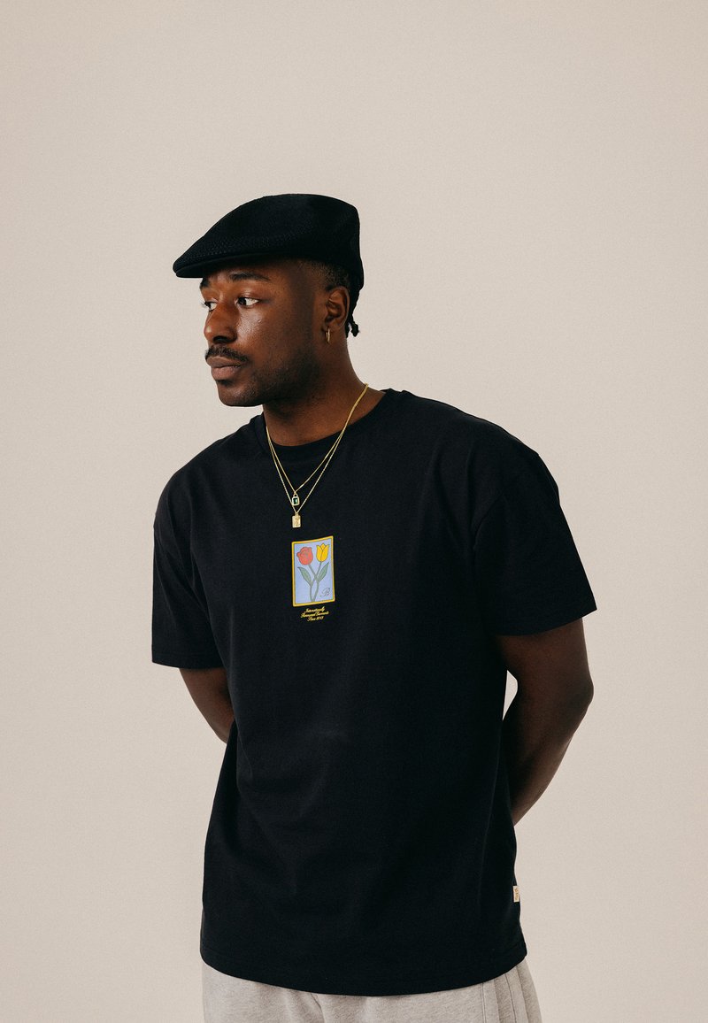 Homme portant un béret noir, un t-shirt noir avec un motif de tulipe, des colliers en or et un pantalon gris clair, regardant vers sa gauche sur un fond uni.