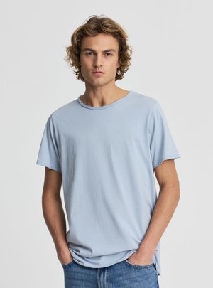 Giovane uomo con capelli biondi ricci che indossa una maglietta celeste a maniche corte e jeans blu, in piedi con le mani nelle tasche.