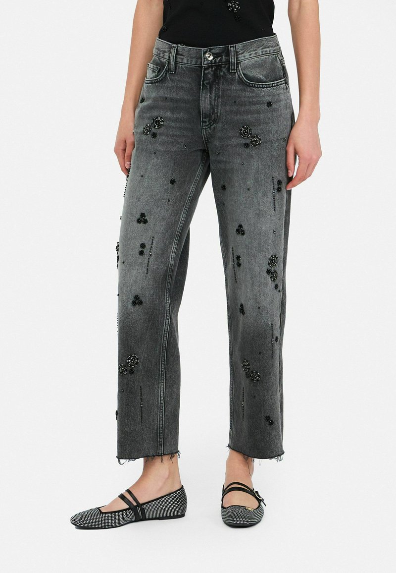 Zwarte denim jeans met een verweerd uiterlijk en gestructureerde zwarte versieringen, gecombineerd met lage zwarte platte schoenen met rhinestone-accenten.