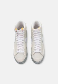 Une paire de baskets montantes Nike beiges avec des lacets blancs, des logos swoosh blancs et des semelles texturées grises, vue de dessus sur fond blanc.