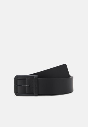 Ceinture en cuir noir roulée avec une boucle métallique rectangulaire noire mate sur un fond clair uni.