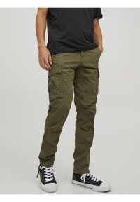 Olive zielone spodnie bojówki z wieloma bocznymi kieszeniami, wykonane z bawełny, o zwężanym kroju nogawki i regulowanych mankietach. Zestawione z czarnymi trampkami.