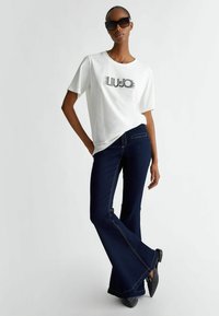 T-shirt bianco a maniche corte con logo "LIU JO" in nero, abbinato a jeans a zampa d'elefante blu scuro con cuciture gialle e una texture liscia.