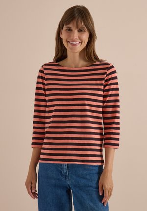 Smilende kvinde med brunt hår iført en pink og sort stribet langærmet top og blå jeans, stående mod en ensfarvet beige baggrund.