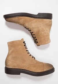 Botas de ante beige con cordones y suelas negras, dispuestas una al lado de la otra sobre un fondo blanco, mostrando los costados exteriores y los detalles de costura.