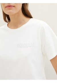 Camiseta blanca de algodón con cuello redondo y mangas cortas, con detalle de texto impreso en azul claro en la zona del pecho izquierdo.