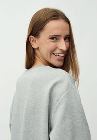 Grauer Pullover mit lässiger Passform, rundem Halsausschnitt und weicher Textur. Das Modell hat lange Haare und trägt kleine Steckerohrringe.