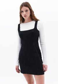 Schwarzes Denim-Kleid mit quadratischem Ausschnitt und breiten Trägern, figurbetonte Silhouette und einer subtilen Textur. Getragen über einem weißen Langarmshirt.