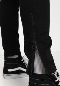Jeans negros con cremalleras laterales, corte ajustado y un acabado texturizado, combinados con zapatillas negras con acentos blancos y cordones texturizados.