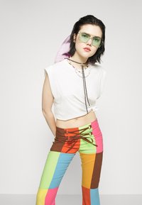 Top curto branco com detalhe em nó, combinado com umas calças elásticas multicoloridas com um design de patchwork e detalhes de amarração. Óculos de sol verdes usados.