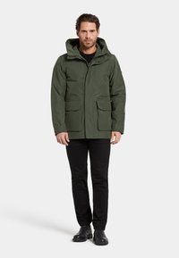 Olivgrön vattentät jacka med huva, två framfickor och dragkedja, parat med svarta byxor och stövlar.