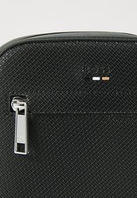 Borsa nera con motivo, dalla superficie texturizzata, dotata di una cerniera argentata e di un logo nero con dettagli bianchi e dorati sopra la cerniera.
