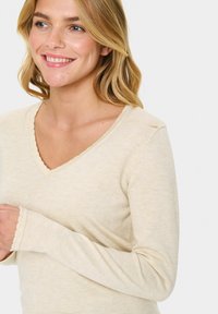 Beidzis ilgajām piedurknēm tops ar V veida kakla izgriezumu un viļņotām malām. Audums ir mīksts, ar teksturētu izskatu, piemērots ikdienas valkāšanai.